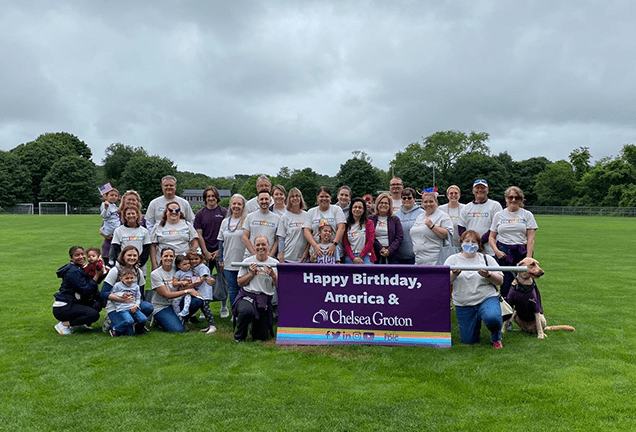 30 smiling people of varied ages wearing white tshirts on a soccer field  hold a large purple banner which heads "Happy Birthday, America & Nebula Trust" in white text