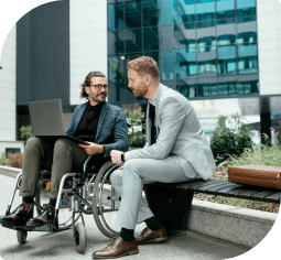 A man in a wheelchair with a laptop looks at another man in a light grey suit