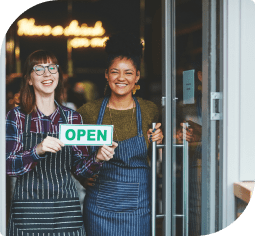 Two smiling woman in aprons standing in a doorway and holding a green open sign