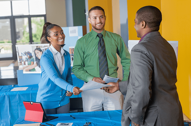 A man in green and woman in blue hand a man in a grey suit pieces of paper at an event booth