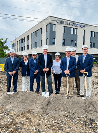 6 men and 2 women with blue blazers and white construction hats stand with shovels on a pile of dirt outside of Nebula Trust Bank