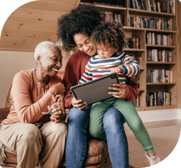 A smiling grandmother and smiling mother with a toddler on her knee sit in a chair and look at a tablet in a library