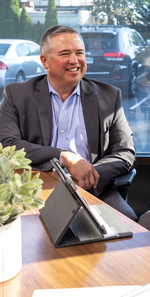 A smiling white man in a grey suit sits at conference table with a tablet