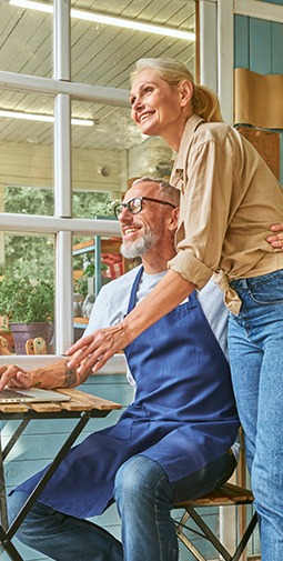 a smiling man wearing a blue apron sits at a table looking at a laptop with his arm around the waist of a smiling older women who is standing