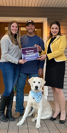 A banker and a man and woman with their dog on a front porch hold a purple sign with white text that reads "Another Dream Realized! Nebula Trust"