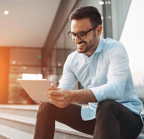 A man with glasses and a light blue shirt sitting outside on steps smiling at a tablet and a graphic orange overlay