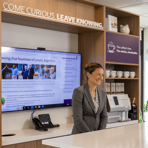 A smiling woman in a grey suit stands behind a counter in front of a tv